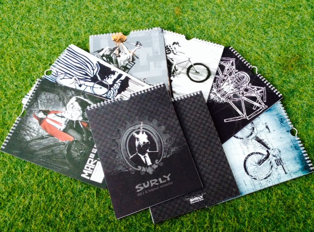 Downward view of 2013 Surly Bikes calendars, opened to different pages, laying on top of each other, on artificial grass