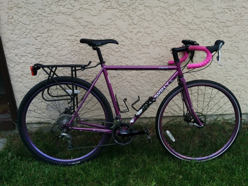 Right side view of a purple Surly Straggler bike parked on grass, leaning against a tan stucco wall