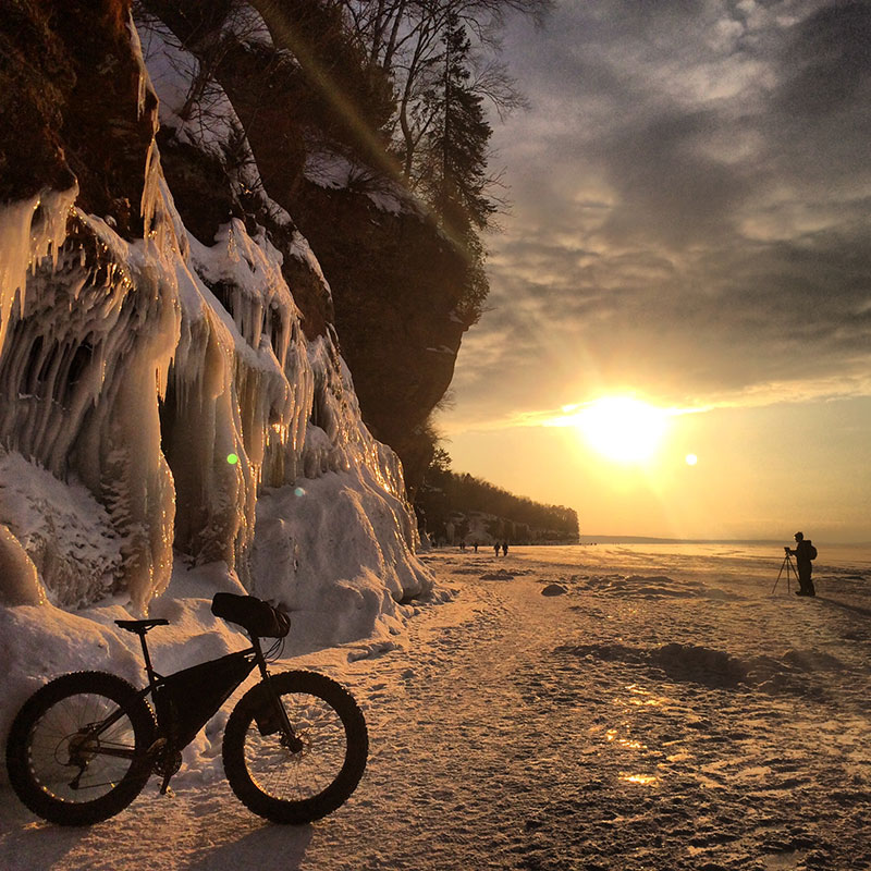 Right side view of a Surly fat bike, next to a cliff wall with icicles, on a frozen lakeshore with the sun shining above