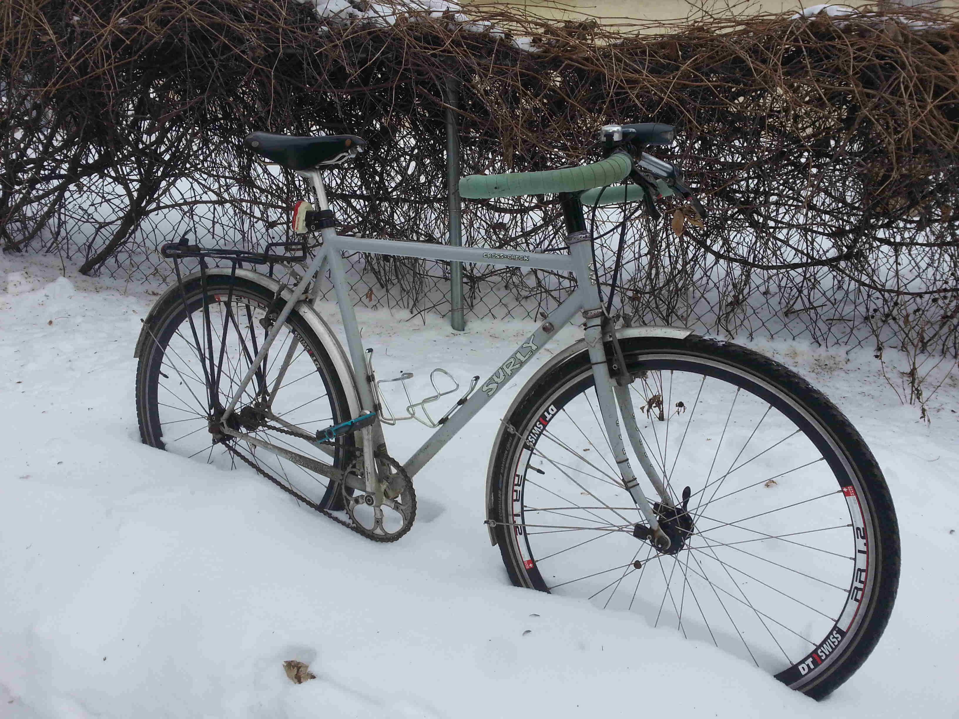 Right side view of a light blue Surly Cross Check bike in deep snow, next to a chain link fence with vines on it