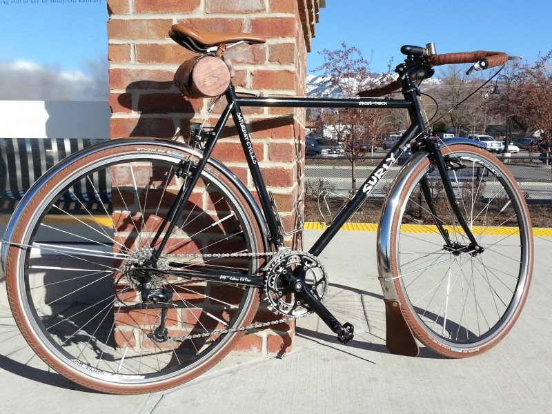 Right side view of a black Surly Cross Check bike, parked on a sidewalk, leaning against a red brick support