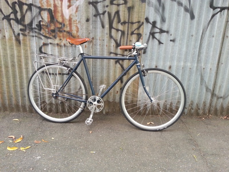 Right side view of a bluish gray Surly bike with white rims, leaning against a steel wall with graffiti on it