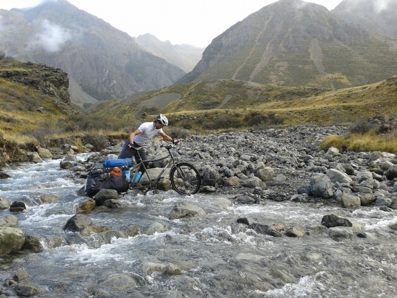 Right side view of a Surly Big Dummy bike, with a cyclist pushing it across a rocky stream in the mountains