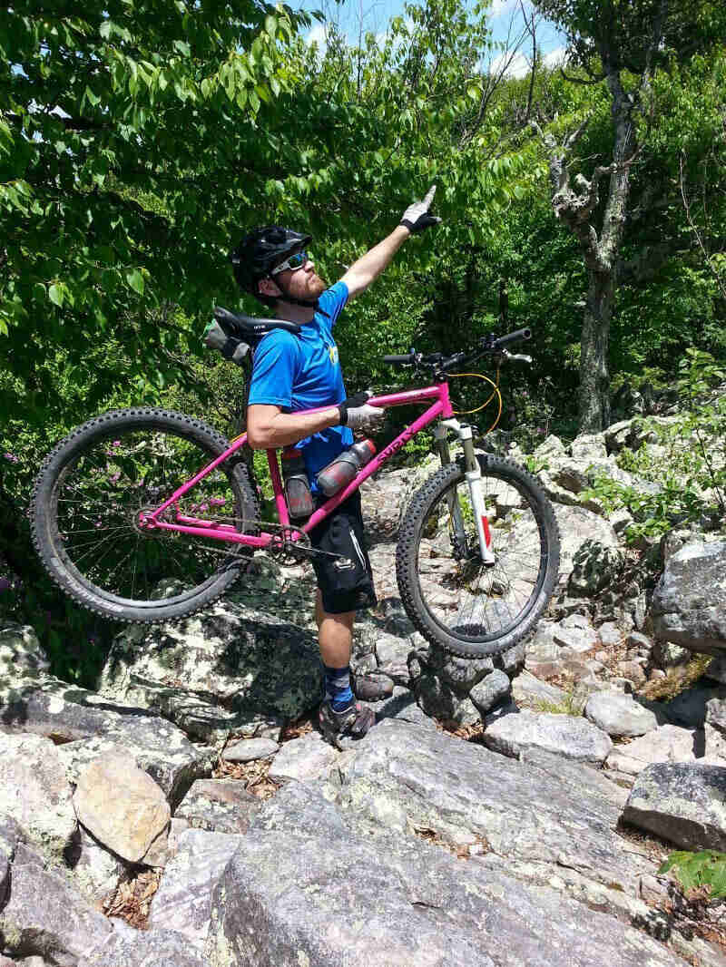 Right side view of a cyclist pointing up into trees, while carrying a pink Surly bike over large rocks