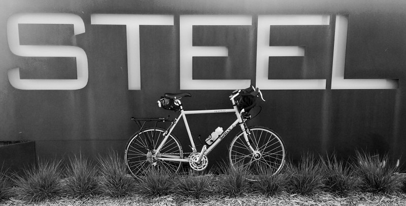 Right side view of a Surly bike, standing in weeds against a wall with, STEEL, spelled in large lettering