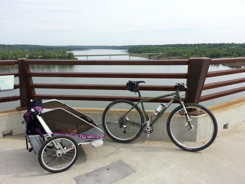 Right side view of an olive drab Surly Ogre bike with a child carrier trailer, on a rail of a bridge with a river below