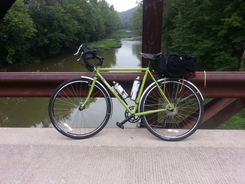 Left side view of a lime Surly bike, parked along a rail on a cement bridge, above a river with trees on the sides