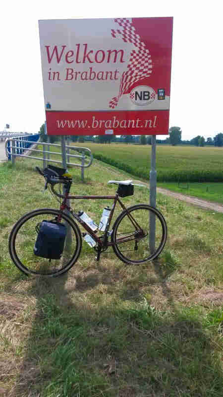 Left side view of a bike with front saddlebags, parked on grass, against a Welkom in Brabant sign