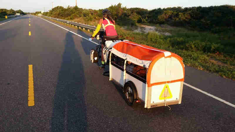 Rear view of a cyclist riding bike down a roadway, with a trailer hitched behind