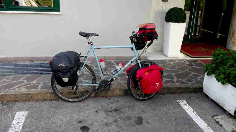 Right side view of a Surly bike, mint, loaded with gear, parked against a curb in front of a white building
