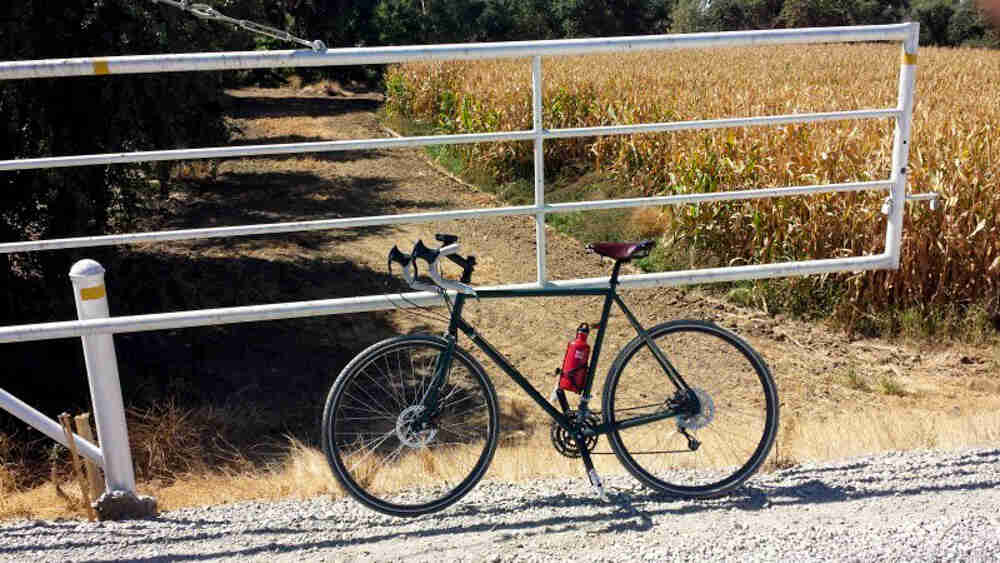 Left side view of a green bike, parked on a gravel road, in front of a white gate, with a cornfield in the background