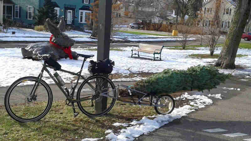 Left side view of a bike and trailer, parked on grass next to a sidewalk, with a big rabbit sculpture on snow behind