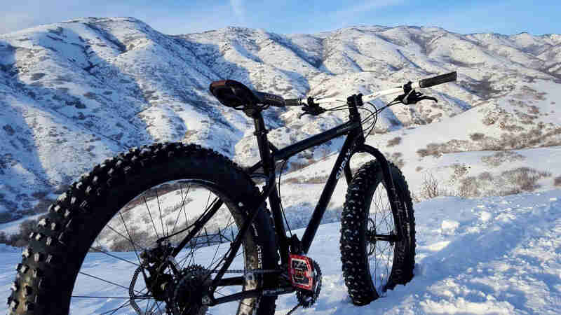 Rear, right side view of a black Surly fat parked in deep snow on a hill, with mountains in the background