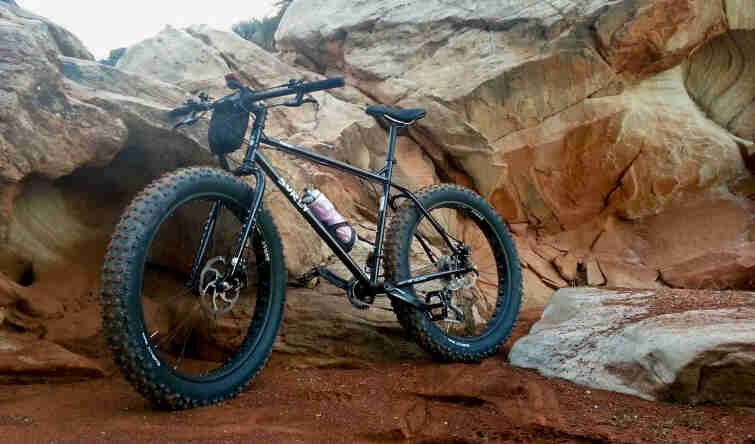 Left side view of a black Surly fat bike, parked on red gravel, in front of a mountain boulder walls