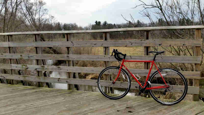 Left side view of a red Surly bike parked, on a bridge over a stream, along the handrail, with trees in the background