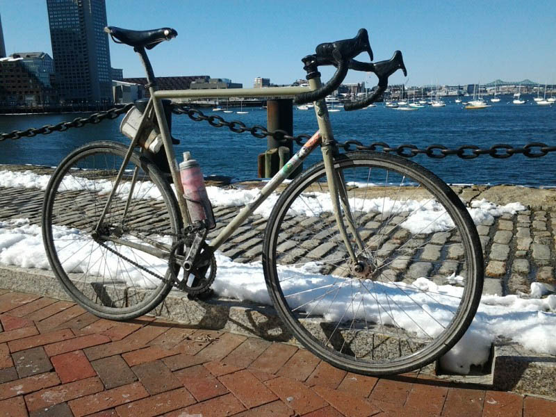 Right side view of a Surly Steamroller bike, gray, parked against curb on a brick street, with a bay in the background