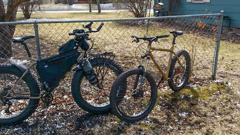 Side view of an olive Surly fat bike with a frame bag, facing another bike, parked against a fence in a yard