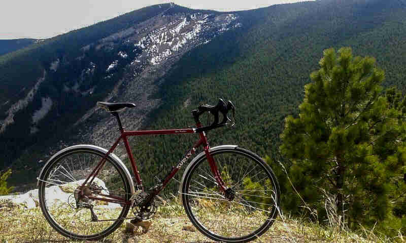 Right side view of a red Surly bike with fenders, parked on a hilltop, with tree covered mountain in the background