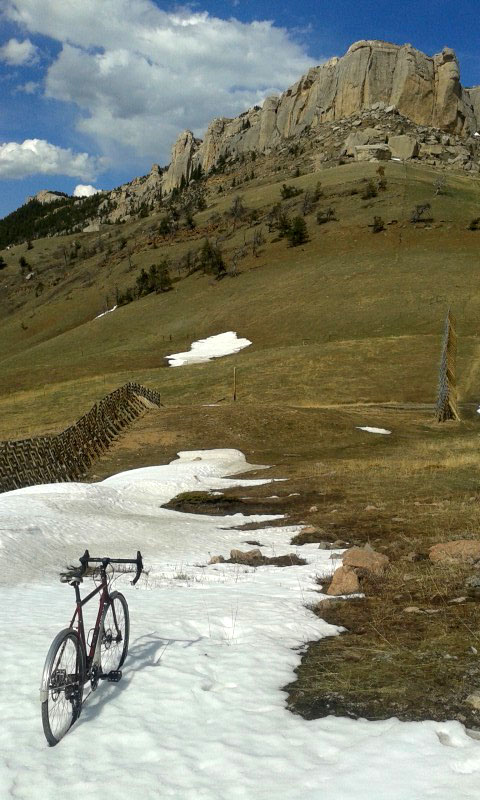 Rear view of bike parked in a patch snow, with a grassy hill and a butte in the background