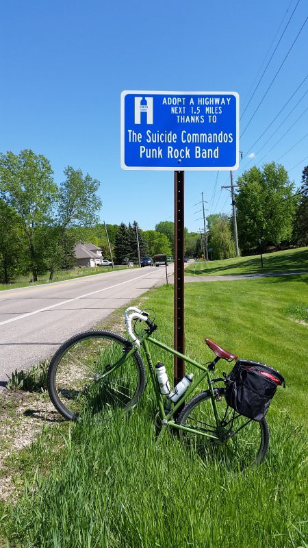 Left side view of green bike parked facing upward, in a grassy road ditch, in front of an Adopt a Highway sign