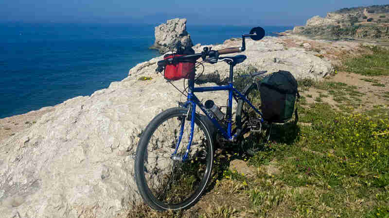 Front left view of a blue Surly bike, parked on top of a rock cliff, above the ocean shown below in the background