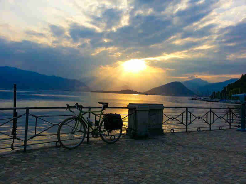 Right side view of a bike, parked along a rail on a stone platform above a lake, with sun shining through the clouds