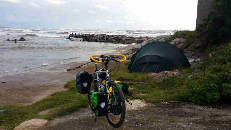 Front view of a Surly bike, loaded with gear, parked on a seashore, with a green tent and the sea in the background