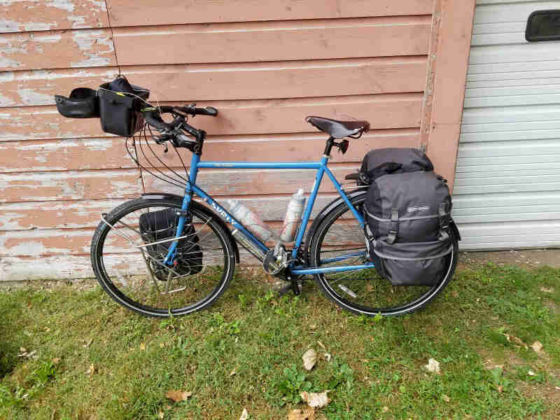 Left side view of a blue Surly bike, loaded with gear, parked in the grass, against a wood siding wall