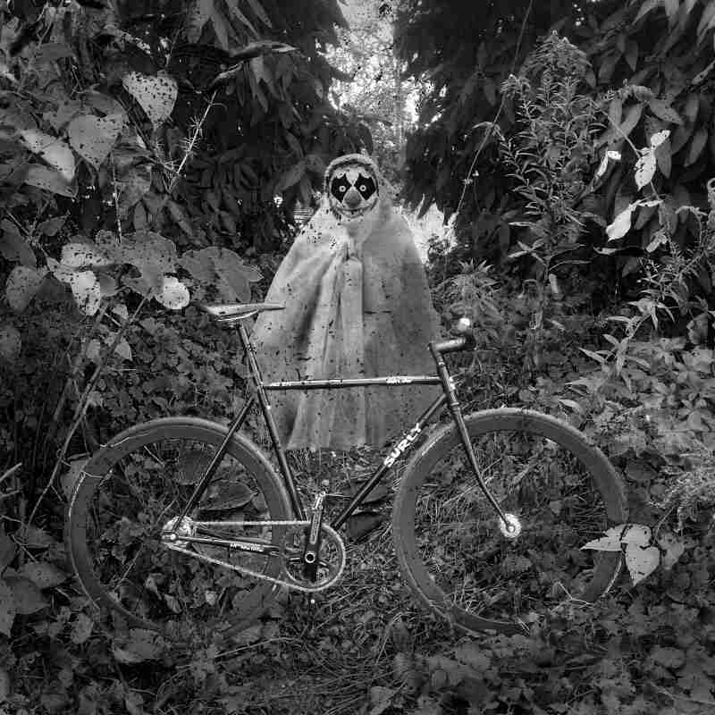 Right profile of a Surly bike parked in thick weeds, with a clown ghost standing behind - black & white image