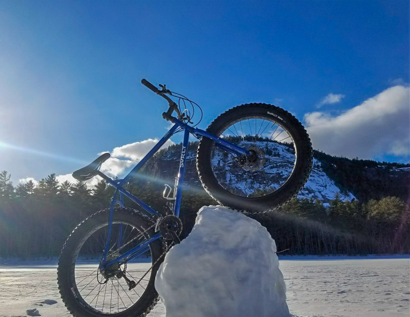 Right side view of a blue Surly fat bike, on a frozen lake, with it's front wheel propped up on a chunk of snow