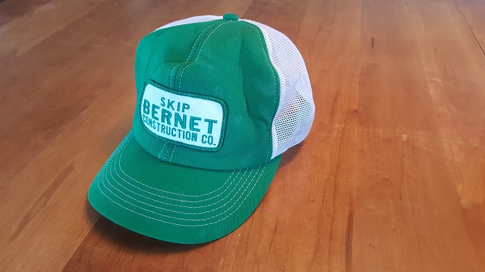 A green trucker hat with a Skip Bernet Construction patch on the front, set on a wood plank floor