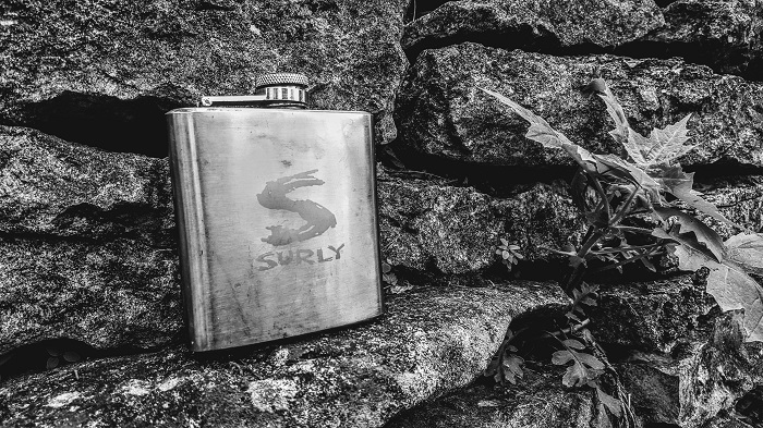 A silver Surly whisky flask sitting in front of a rock wall, black and white rendering
