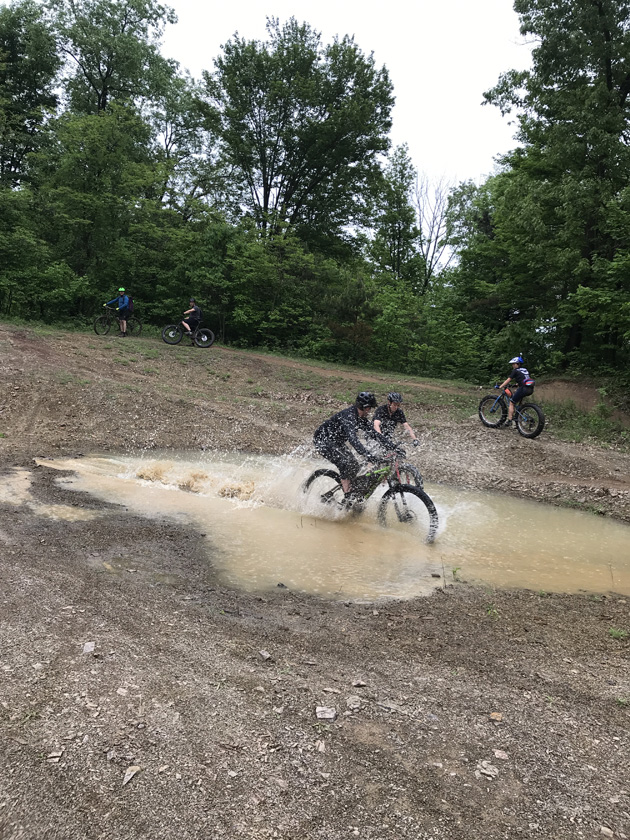 Cyclists ride through dirt and mud in a field with trees in the background