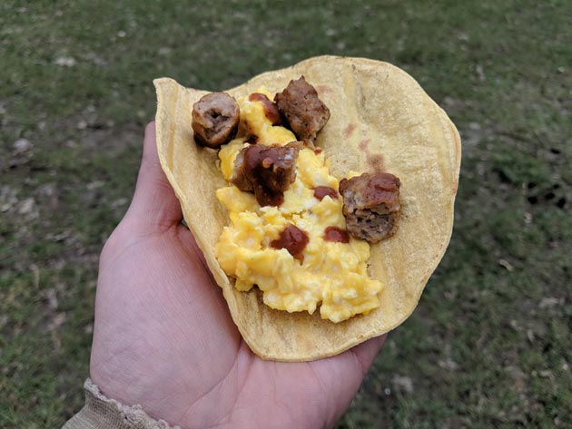 Downward view of a hand holding a tortilla with sausage and eggs inside and grass on the ground in the background