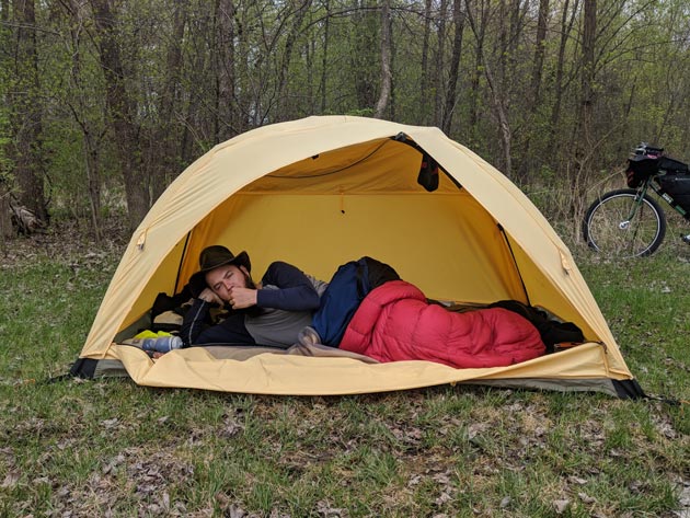 Person laying in a red sleeping bag inside a yellow dome tent on a grass field with a bike and trees in the background