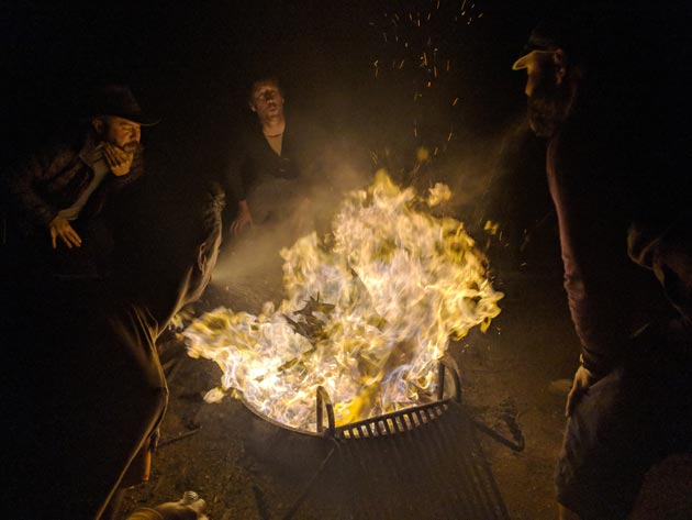 A raging fire inside a campfire ring with 2 people sitting behind at night