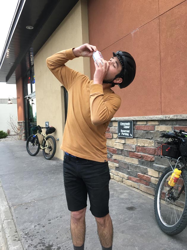 Cyclist shotgunning a beer in front of a building with a bike behind leaning on a stone wall