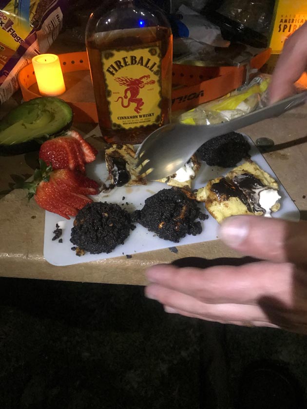 A hand holding a spork above a small cutting board with food on a table with a bottle of Fireball whisky behind