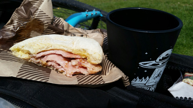 A black cup and a half eaten sandwich siting on a black bike bag