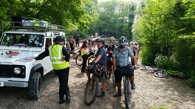 A group of cyclist with their bikes stand on a gravel lot in the trees next to a white SUV 