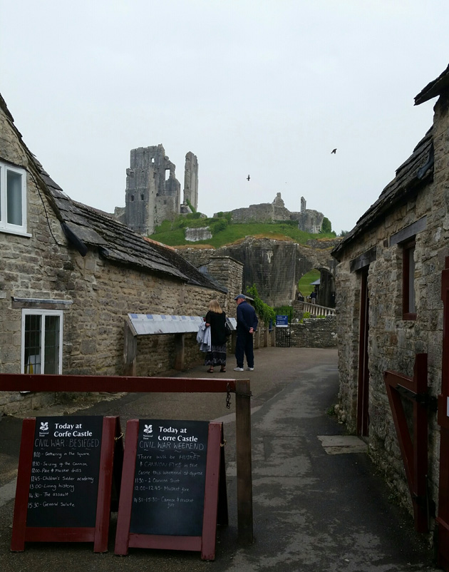 Two persons walking on a narrow road between stone buildings with Corfe castle in the background up the hill