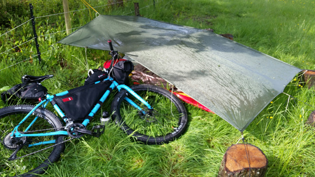 Surly bike, turquoise, loaded with gear, laying on the left side in the grass next to a gray tarp covering equipment 