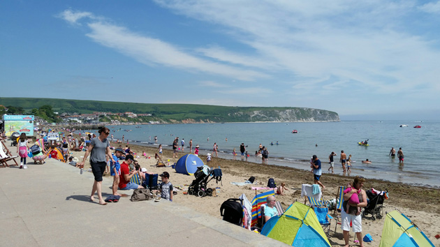 People on a beach and in the water with grassy rock land across the bay on a sunny day with few clouds