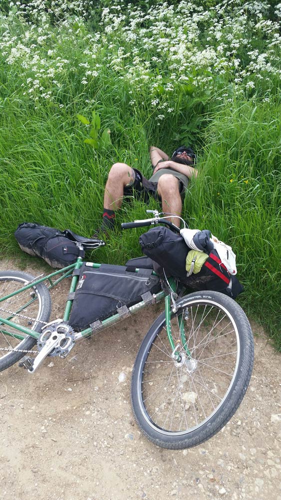 A cyclist laying down in tall, green grass and their green bike lying on it's left on top of gravel