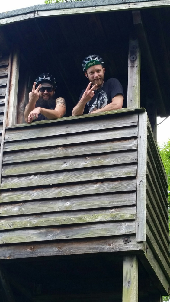 Two people standing next to each holding 2 finger up while in an opening of a wood building on stilts
