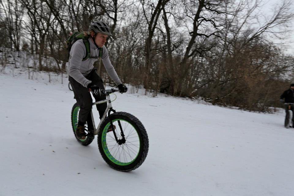 Right side view of a cyclist riding a white Surly fat bike down a snowy hill, with trees in the background