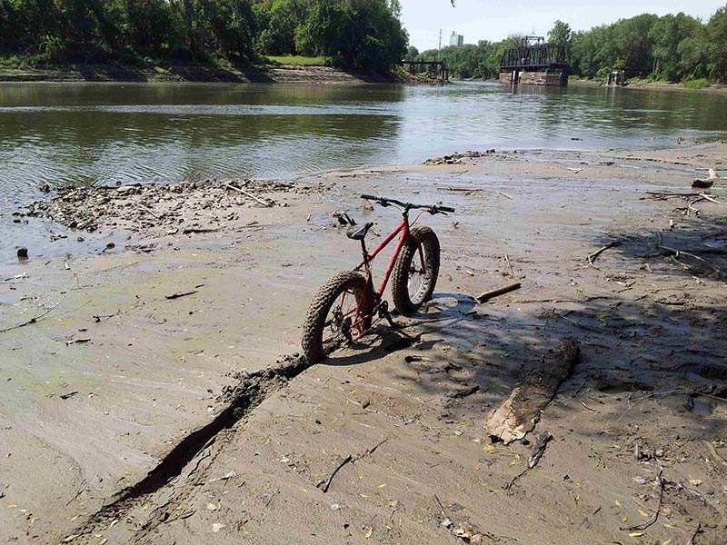 Rear, right side view of a red Surly Moonlander fat bike, standing in deep mud, on a flat river bank
