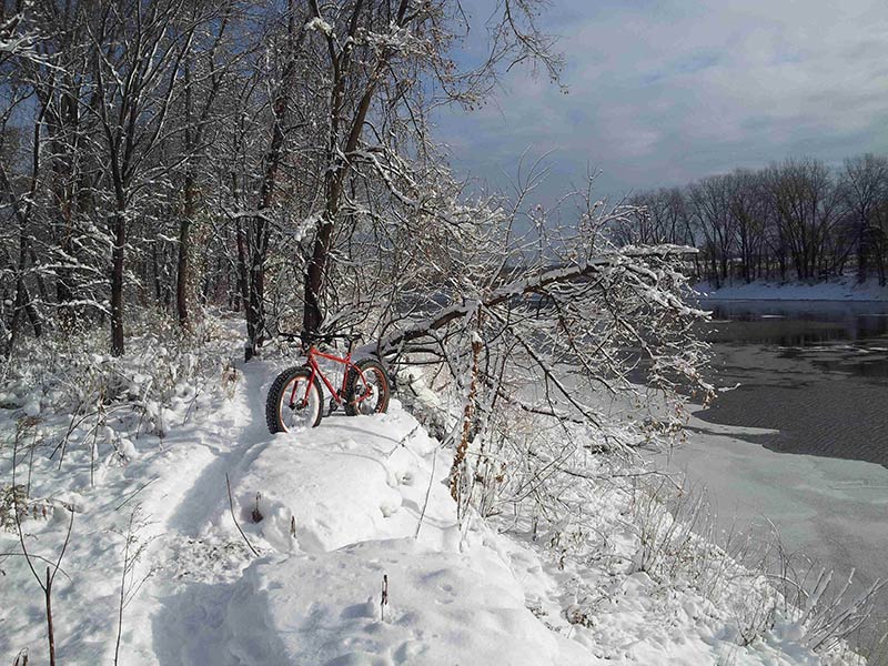 Front, left side view of a red Surly Moonlander fat bike, standing in deep snow, on a trail next to a river