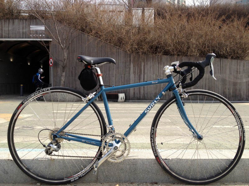 Right side view of Surly Pacer bike, blue, against a curb of a roadway, with a tunnel and cement wall in the background