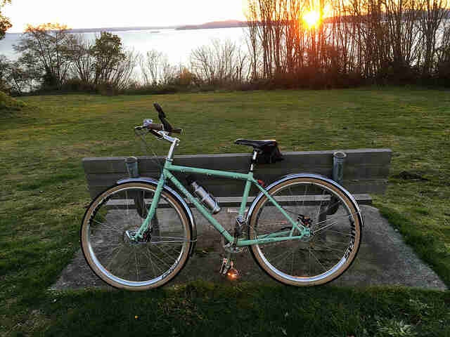 Left side view of a mint Surly bike, leaning against the back of a park bench,  with a lake in the background